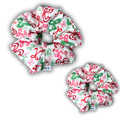 House of Woof Christmas scrunchies featuring a festive red, green, and pink ribbon pattern on white fabric. Handmade holiday hair accessories for dog lovers and matching pup sets.