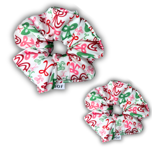 House of Woof Christmas scrunchies featuring a festive red, green, and pink ribbon pattern on white fabric. Handmade holiday hair accessories for dog lovers and matching pup sets.